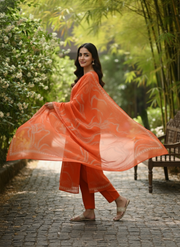 Woman in an orange traditional outfit standing in a garden with greenery.