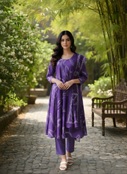 Woman in a purple outfit standing in a garden with greenery and a wooden bench.
