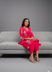 Woman in a bright pink dress with floral embroidery sitting on a gray couch against a gray background